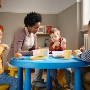 Early years practitioner sitting with toddlers at a low table at lunchtime