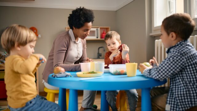 Early years practitioner sitting with toddlers at a low table at lunchtime